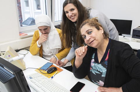 Zwei Frauen sitzen am Computer, eine dritte beugt sich von hinten ber sie.